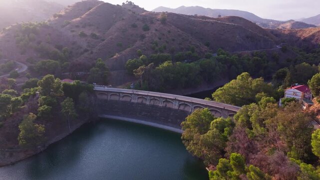 Drone footage of Embalse del Limonero dam near Malaga, Spain, tracking a historic arched bridge over turquoise reservoir waters amid sunlit hills.