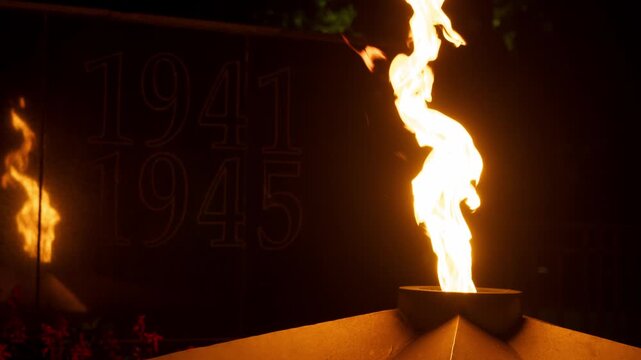 Bright eternal flame illuminates memorial displaying dates 1941 1945 at night. Monument commemorates fallen soldiers, wartime sacrifice, victory, and historical remembrance.