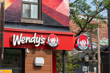 Obraz premium Toronto, ON, CANADA - Aug 8 2024 : Exterior of a Wendy's restaurant with the red logo signage and Wendy mascot circle signs mounted on a red awning below a brick building facade.