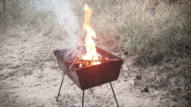 Dry wood burns with bright orange flames inside a portable metal grill on a sandy outdoor field. Concept of barbecue preparation, camping lifestyle, outdoor cooking, and weekend leisure.