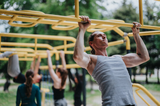 A muscular man performs pull-ups on yellow monkey bars during an outdoor workout. Other people train in the background at a park fitness area.
