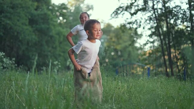 Boy leads sack race jumping in summer. Girl follows close in outdoor game. Children enjoy playful competition. Kid bounces ahead. Summer game brings joy to every child. Girl and boy love outdoors fun.