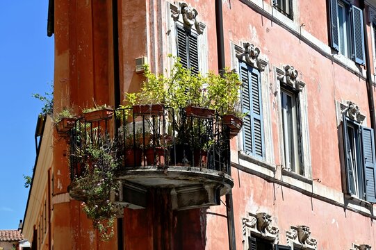 classic old circular metal balcony. black steel picket balustrade and railing. flowers in plastic pots.  deteriorating cantilevered concrete slab. spalling stucco exterior elevation. decorative window
