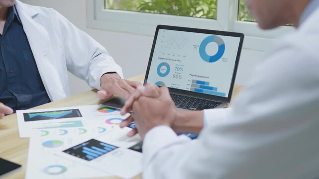 Doctors consulting together and planning treatment in a modern medical office, reviewing healthcare data, charts, and reports on a laptop for clinical decision making and patient care.
