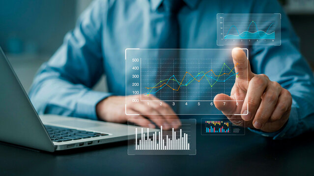 A businessman interacting with a futuristic holographic interface displaying financial charts and data analysis. The user is pointing to a graph on the screen.