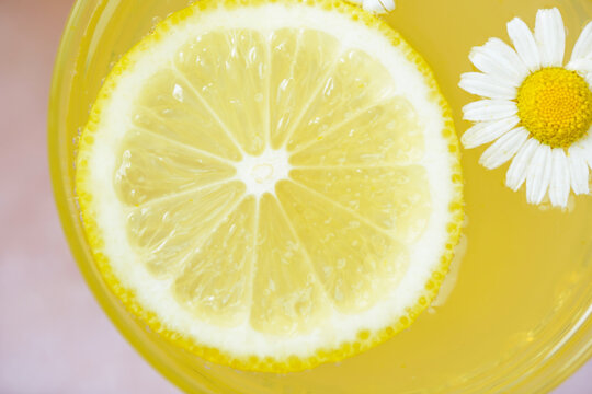 Close-up macro view of a fresh lemon slice floating in a bright yellow citrus drink with a white chamomile flower. Top view. Perfect for healthy drinks, wellness, detox, lemonade, herbal tea