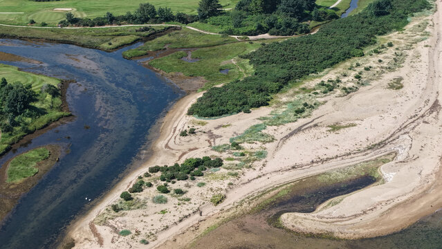 Aerial view river estuary meeting coastal sand dunesRiver estuary winding through sand, beach, and green golf course landscape