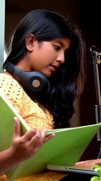 Indian Asian teenage girl studying online at home using a laptop computer while sitting by a bright, airy window in a cozy lavish home, focused on education, exam preparation, learning with dedication