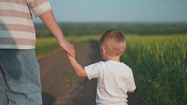 Mother holds child hand on the road. The boy, a lively kid, joins the parent for a summer path walk. Mother talks to child, boy sings, kid skips by the field. Parent and child love a summer road day
