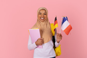Smiling young Black Muslim student in beige hijab holding international flags and notebooks with yellow backpack, representing global education on a pink background © Anastasiya