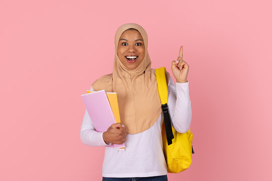 Excited young Black Muslim female student in beige hijab holding notebooks and raising one finger, having idea while carrying yellow backpack on pink studio background