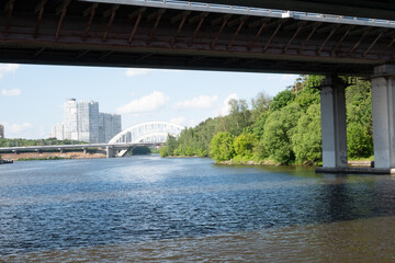 Horizontal view from beneath a dark bridge frame, looking down a river toward a white arch bridge and modern white residential towers. Great for city landscapes and travel photography. © Margarita