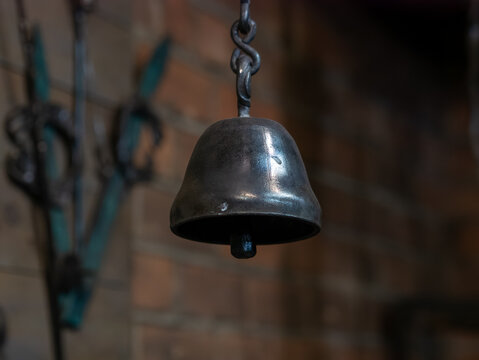 A close-up shot of a handcrafted dark metal bell hanging from a twisted chain against a blurred brick wall inside a rustic workshop.