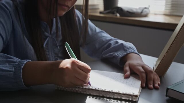 Tilt up view of teenage girl writing in spiral notebook at desk, focusing on schoolwork. Footage for educational marketing, tutoring services, and online learning advertisements