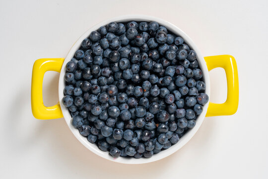 Fresh ripe blueberries piled high in white ceramic bowl with bright yellow handles, closeup. Flat lay of organic summer berries isolated on clean white backdrop. Perfect for healthy eating concepts