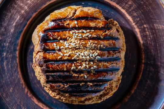 Golden brown homemade pastry topped with sesame seeds and egg wash glaze on rustic clay plate, closeup. Freshly baked artisan bun captured from above. Ideal for bakery marketing and food concepts