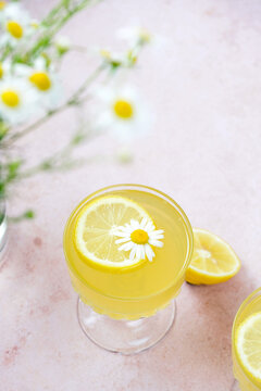 Yellow drink made with lemon sits in a glass. It features a flower and a lemon slice on top. Surrounding the drink are fresh flowers. The setting is bright and cheerful.