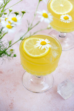 Yellow drink made with lemon sits in a glass. It features a flower and a lemon slice on top. Surrounding the drink are fresh flowers. The setting is bright and cheerful.