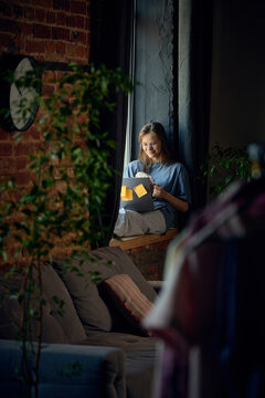 Smiling young woman using laptop and holding coffee in cozy window workspace. Concept of client communication, remote meetings, online appointments and flexible customer service.