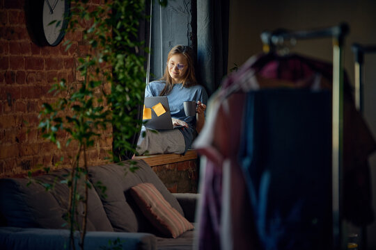 Relaxed woman combining laptop work with coffee break beside apartment window. Concept of content planning, social media management, digital marketing and creative business workflow.