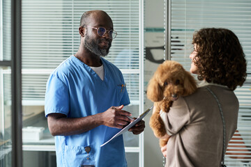 Medium shot of Black male veterinarian consulting female pet owner, holding clipboard while examining poodle in clinic. Ideal for veterinary care, pet health, animal service marketing © Mediaphotos