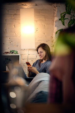 Woman using smartphone and laptop during evening rest at home. Concept of digital entertainment, streaming subscriptions and leisure content consumption.