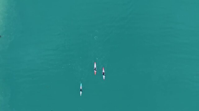A high altitude view looking straight down on three kayakers zipping across calm, beautifully colored waters of the Pacific Ocean.