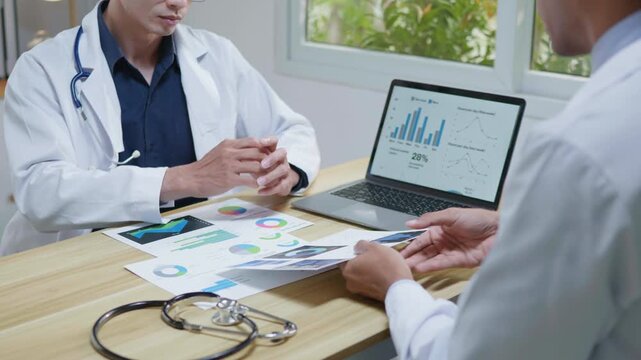 Doctors consulting together and planning treatment in a modern medical office, reviewing healthcare data, charts, and reports on a laptop for clinical decision making and patient care.