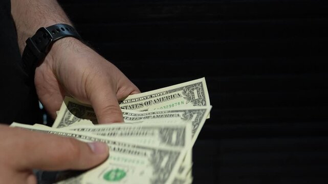 A mans hands carefully count several one dollar US banknotes against a dark backdrop. Cinematic lighting highlights the texture of the currency.