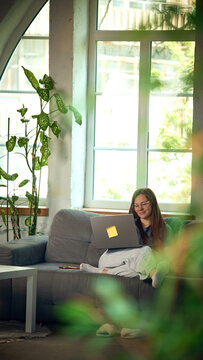 Woman working with laptop while sitting comfortably on sofa near bright window. Concept of online learning, professional development and self-paced digital education.