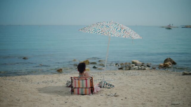 Woman is sitting in a beach chair reading a book, finding shade under an umbrella at a tranquil sandy beach