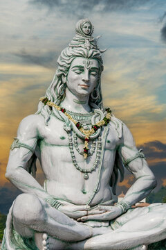 Majestic statue of Hindu deity Lord Shiva in deep meditation. Spiritual monument decorated with flower garlands under beautiful dramatic sunset sky. Ideal for yoga, travel, and religious concepts