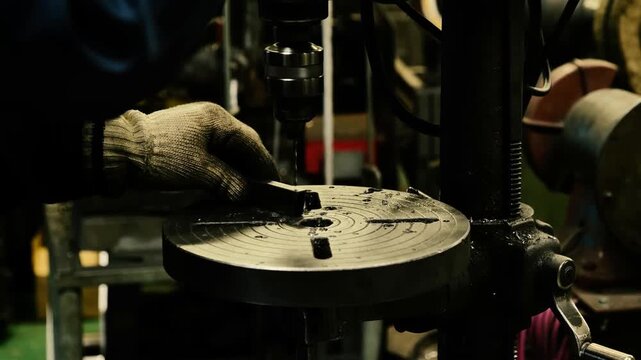 The professional metalworker uses a manual drill press in a machine shop to bore precise holes into a small steel workpiece with industrial tools and specialized equipment.