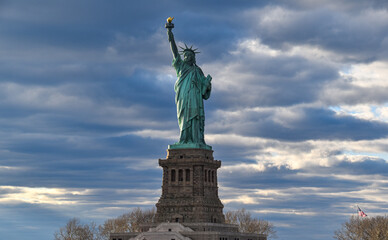 The iconic Statue of Liberty standing proudly in New York Harbor beneath dramatic storm clouds. A striking commercial travel and architecture image showcasing one of America's most famous landmarks. © Gerard