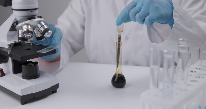 Scientist dispensing dark oil liquid into glass flask beside microscope on laboratory benchtop in chemical lab.