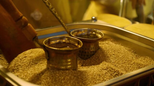 Traditional Turkish coffee brews in copper cezve on hot sand. Street food vendor stirs sand around pot to maintain even heating during brewing.