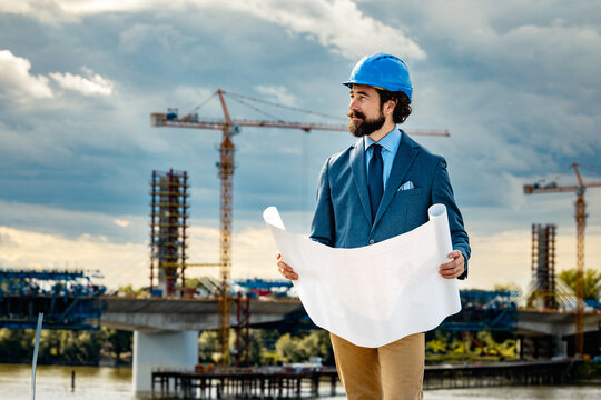 Construction engineer in suit and hard hat reviewing blueprints at bridge building site