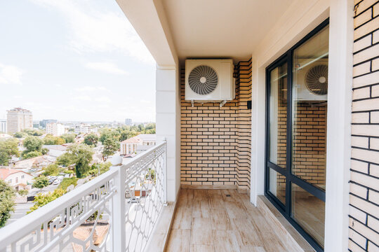 Sunlit balcony with wall mounted air conditioner against beige brick exterior beside sliding glass door, white metal railing, and distant city skyline view from high floor apartment