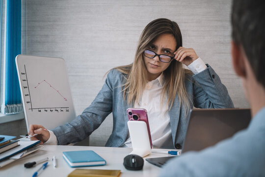 Strict female manager looking disapprovingly at an employee during a business meeting. Serious discussion about poor sales performance, leadership, office management and workplace communication.