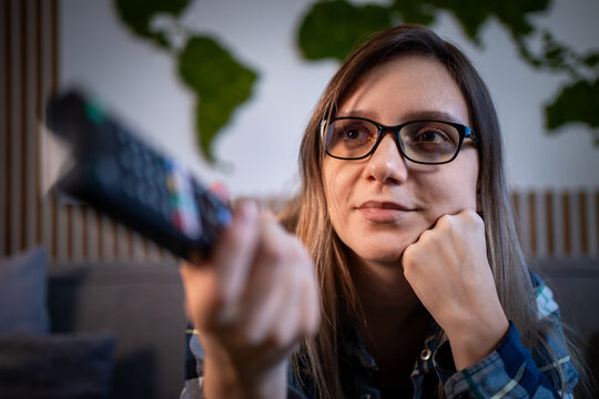 Bored young woman with glasses sitting on sofa and switching TV channels with remote control. Tired of watching television.