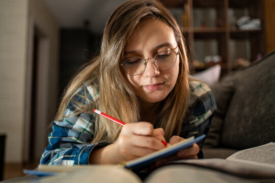Smart student young woman writing in a notebook while lying on a sofa with a stack of books. Home studying, creative writing, and cozy student lifestyle. writing a curriculum.