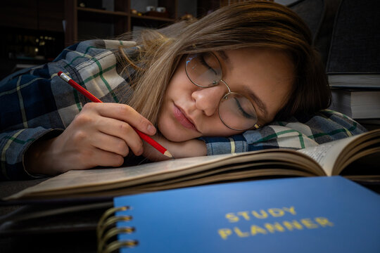 Exhausted female student sleeping on an open book with a red pencil in her hand, next to a blue study planner. Academic burnout, late-night studying, and exam fatigue.