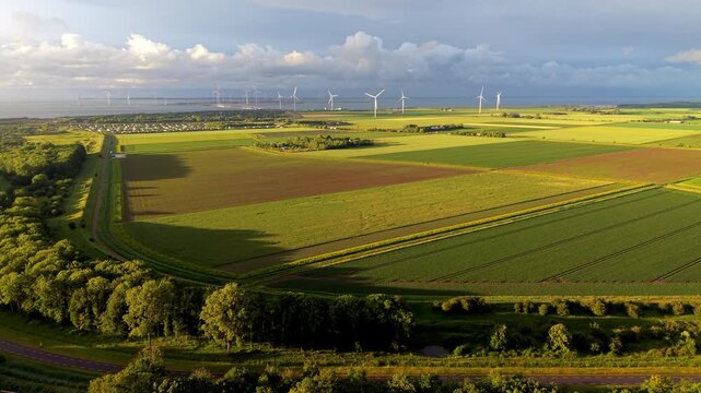 Aerial wide angle panoramic view of vast green agricultural fields and a row of wind turbines on the coast under a dramatic cloudy sky illuminated by warm evening sunset light