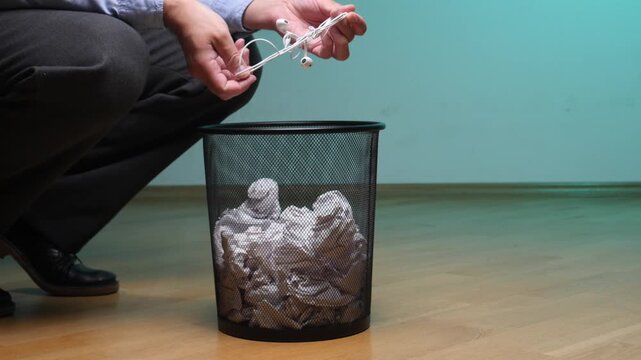 Hand holding tangled white wired earphones, placing them over a black mesh trash bin filled with crumpled paper, symbolizing electronic waste disposal and technological obsolescence