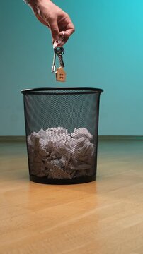 Vertical video hand dropping a set of house keys with a wooden house shaped keychain into a trash can overflowing with crumpled paper, symbolizing loss, foreclosure, and financial failure