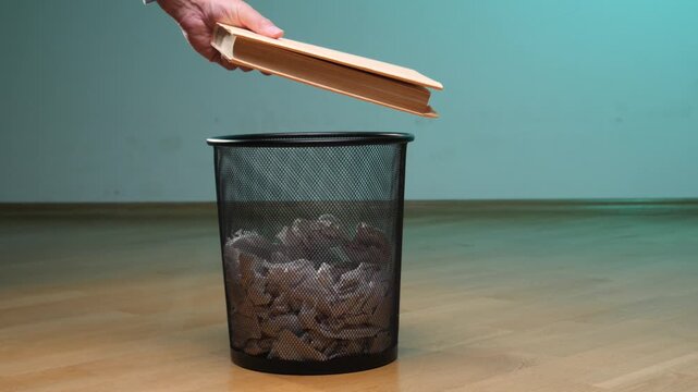 Hand dropping a book into a black mesh waste bin filled with crumpled paper, symbolizing censorship, knowledge rejection, and discarding education