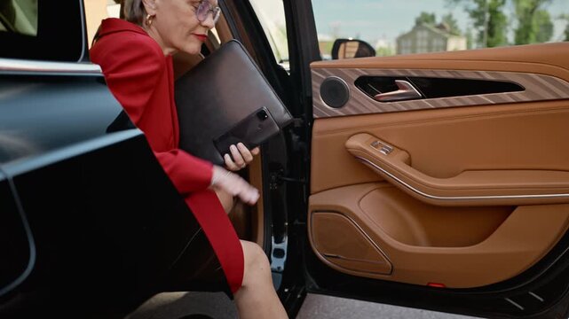 Chauffeur wearing gloves opening luxury car door for middle aged Caucasian businesswoman exiting vehicle holding folder. Clip is ideal for premium corporate transport services