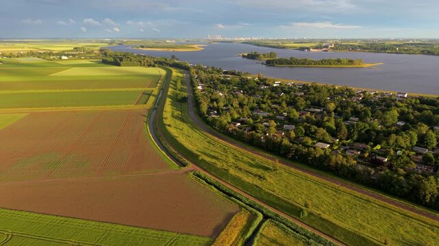 Aerial wide angle panoramic view of a massive green grassy dyke protection wall next to agricultural fields and a cozy lakeside village under a dramatic sunset sky in warm evening light