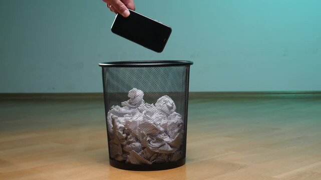 Hand dropping a modern smartphone into a metal mesh trash bin filled with crumpled white paper, symbolizing digital detox, data privacy, and e waste disposal