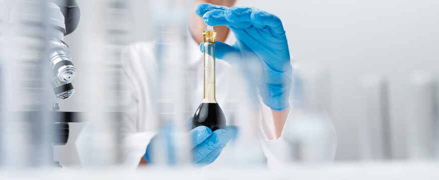 Closeup of gloved hands holding dark oil in glass flask for laboratory analysis and testing background